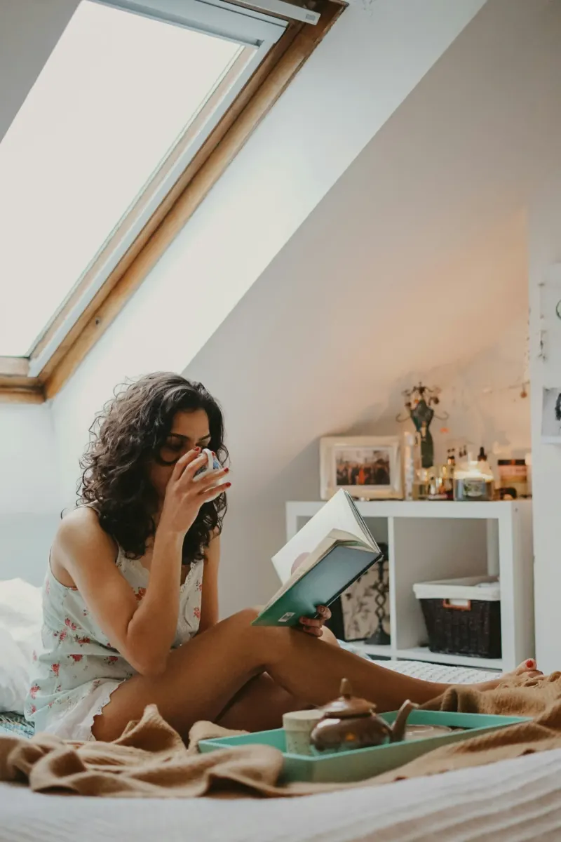 Femme lisant un livre, une tasse à la main, assise sur son lit