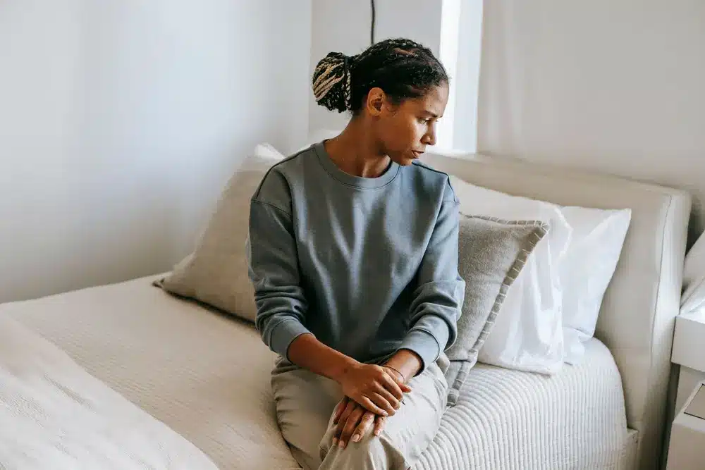 Une jeune femme assise sur un lit blanc, les mains jointes, le regard baissé vers le sol