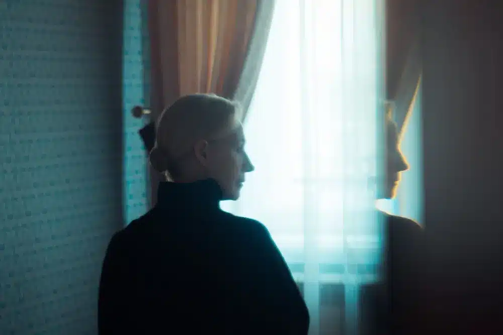 Silhouette d’une femme de profil devant une fenêtre aux rideaux clairs, dans une pièce sombre