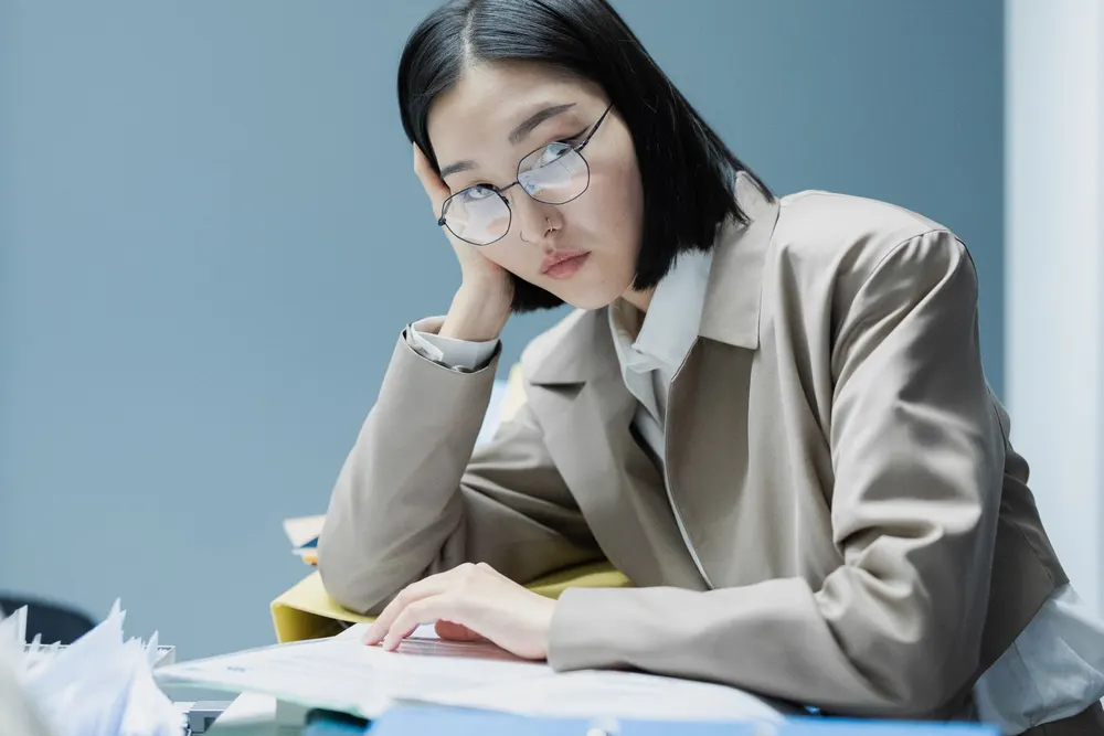 Une femme à lunettes, le menton appuyé sur la main, l’air las, devant des documents posés sur un bureau