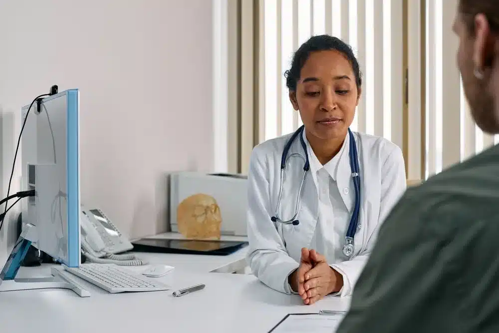 Une médecin en blouse blanche, les mains jointes, écoute un patient pendant une consultation dans son cabinet