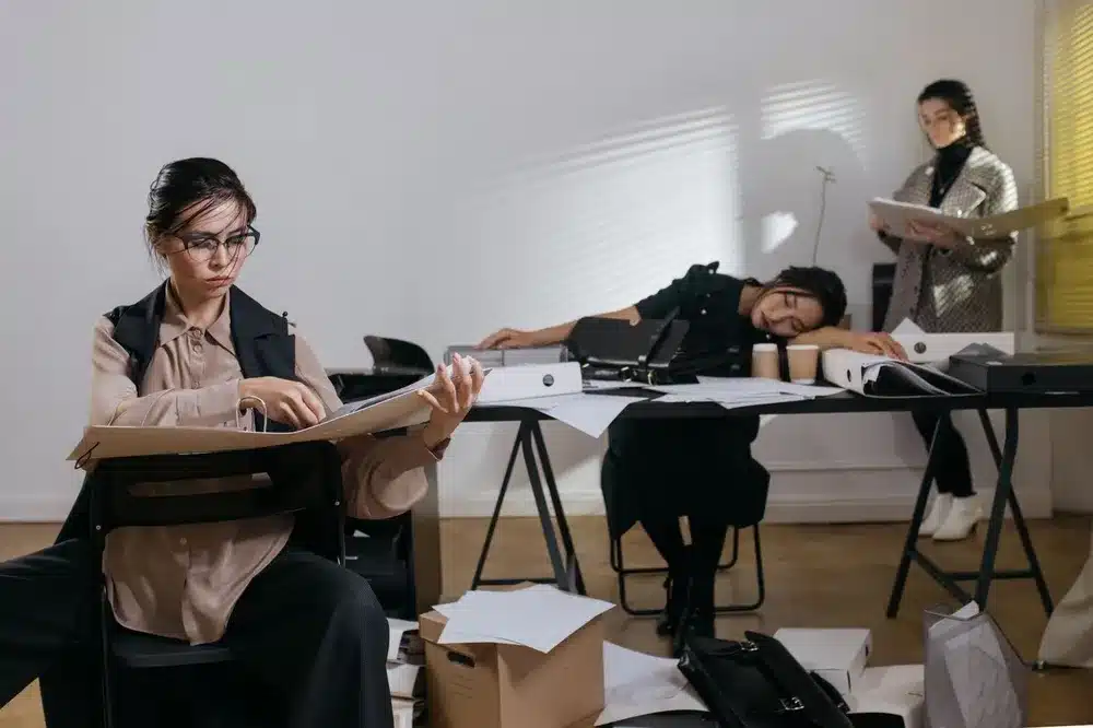 Bureau encombré de classeurs et de feuilles, avec une femme qui lit un dossier au premier plan et une autre qui repose la tête sur la table