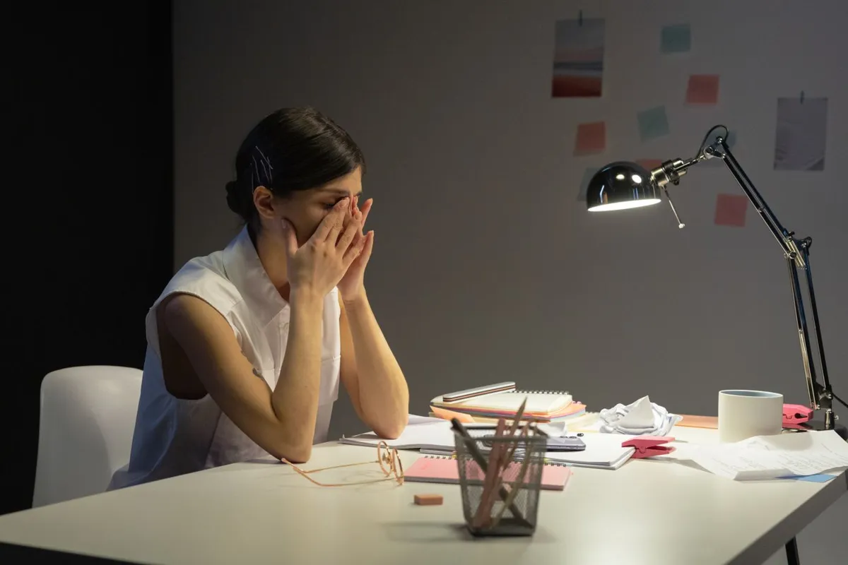Femme assise à son bureau, les mains sur le visage, devant une lampe et des papiers