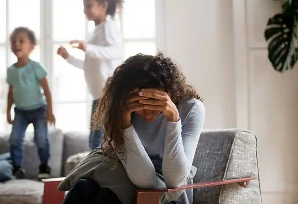 Une femme assise sur un canapé, la tête dans les mains, pendant que deux enfants jouent derrière elle