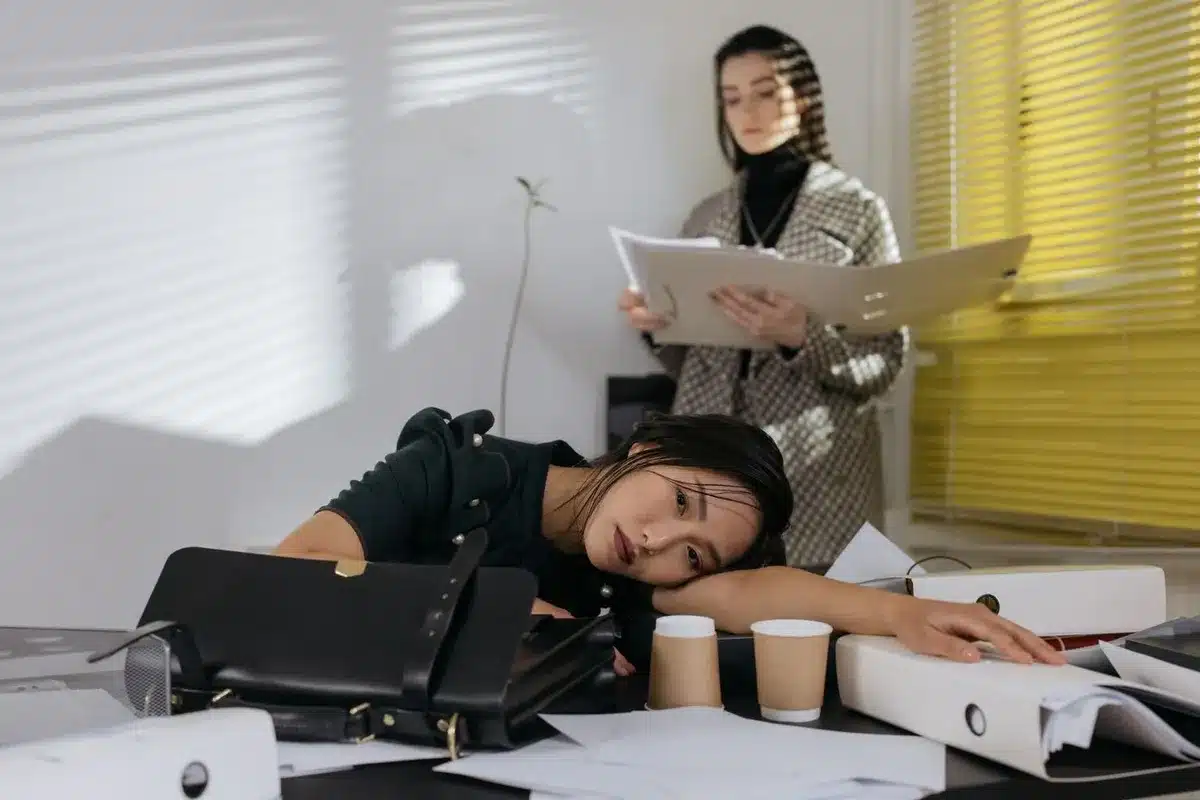 Femme qui repose la tête sur son bureau encombré de classeurs, une collègue debout derrière elle lisant un dossier