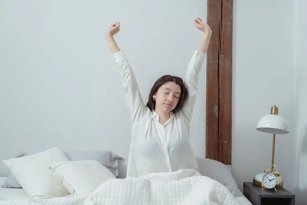 Une femme assise dans son lit s'étire les bras levés au réveil, un réveil posé sur la table de nuit