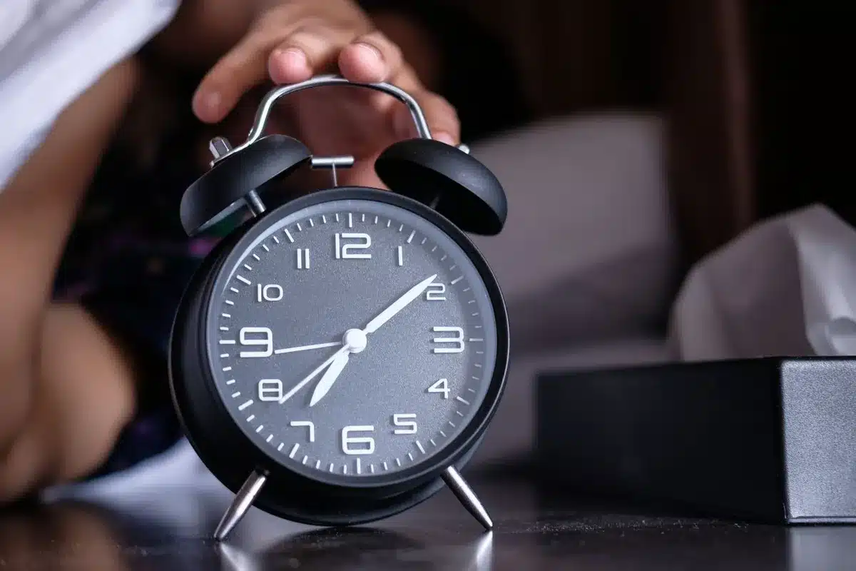 Une main posée sur un réveil à aiguilles noir, au bord d'un lit, au petit matin