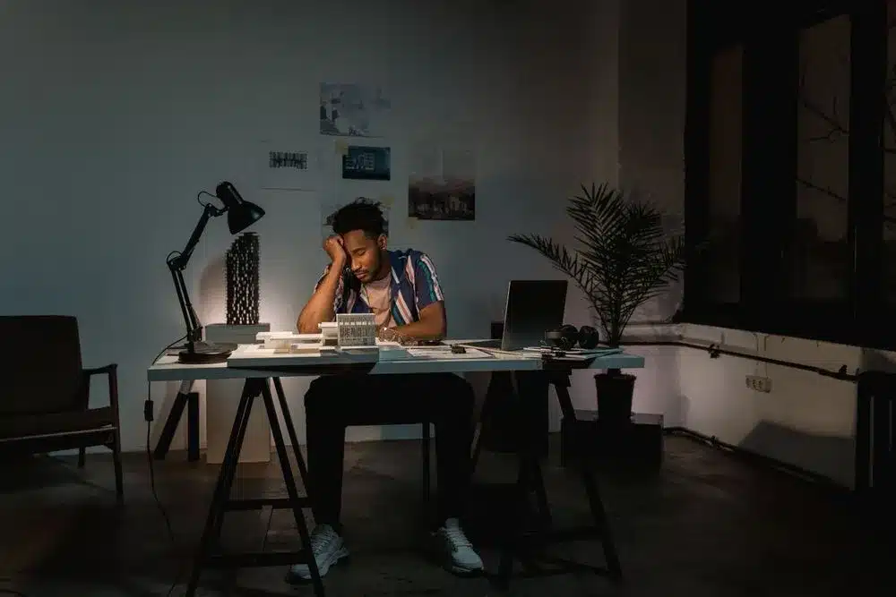 Homme assis à un bureau éclairé par une lampe, la tête appuyée sur la main, dans un atelier sombre